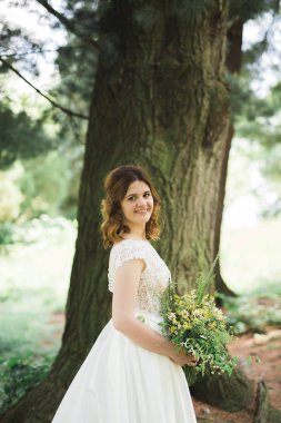 Zarif beyaz elbiseli güzel esmer gelin elinde buket ile güzel ağaçlar poz veriyor.