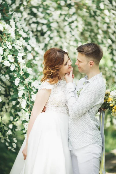 Elegante coppia di sposi felici che passeggiano nel parco il giorno del loro matrimonio con bouquet — Foto Stock