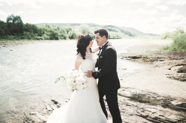 Schönes Hochzeitspaar küsst und umarmt sich am Ufer eines Gebirgsflusses mit Steinen — Stockfoto