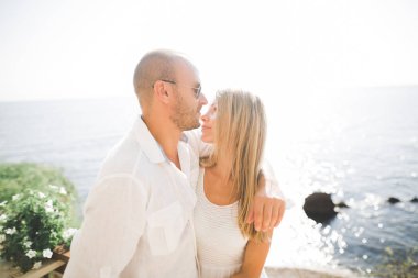 Düğün çifti mavi denizin yanındaki kayalarda öpüşüyor ve sarılıyor.