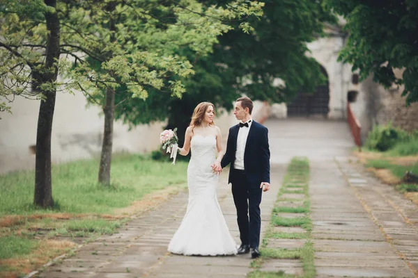 Bella coppia di sposi felici ed eleganti che passeggiano nel parco il giorno del loro matrimonio con bouquet — Foto Stock