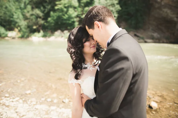 Красивая свадебная пара целуется и обнимается у берега горной реки с камнями — стоковое фото