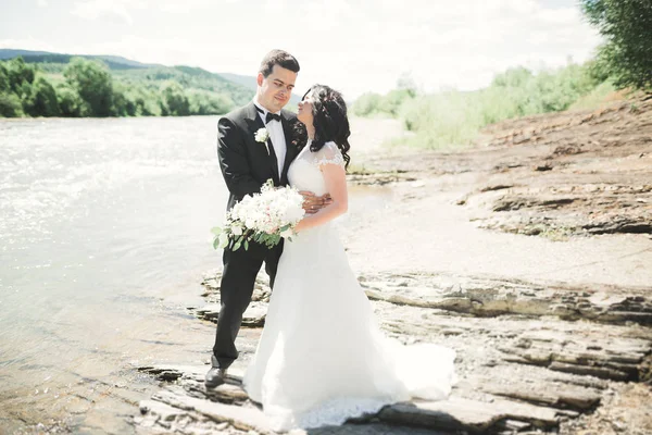 Elegante stilvolle glückliche brünette Braut und wunderschöner Bräutigam vor dem Hintergrund eines wunderschönen Wasserfalls in den Bergen — Stockfoto