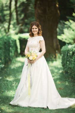 Zarif beyaz elbiseli güzel esmer gelin elinde buket ile güzel ağaçlar poz veriyor.
