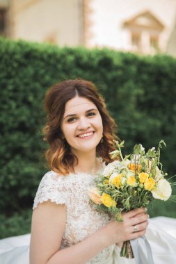 Zarif beyaz elbiseli güzel esmer gelin elinde buket ile güzel ağaçlar poz veriyor.