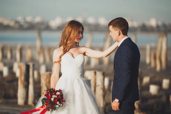 Casal de casamento jovem bonito, noiva e noivo posando perto de postes de madeira no fundo do mar — Fotografia de Stock