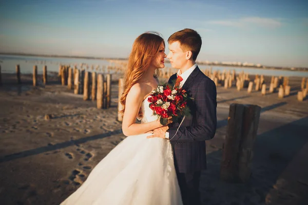 ΓΑΜΟ ζευγάρι, γαμπρός, νύφη με ανθοδέσμη που παρουσιάζουν κοντά στη θάλασσα στο ηλιοβασίλεμα — Φωτογραφία Αρχείου
