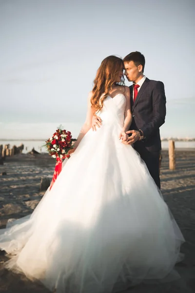 Красивая молодая супружеская пара, невеста и жених позируют возле деревянных столбов на фоне моря — стоковое фото