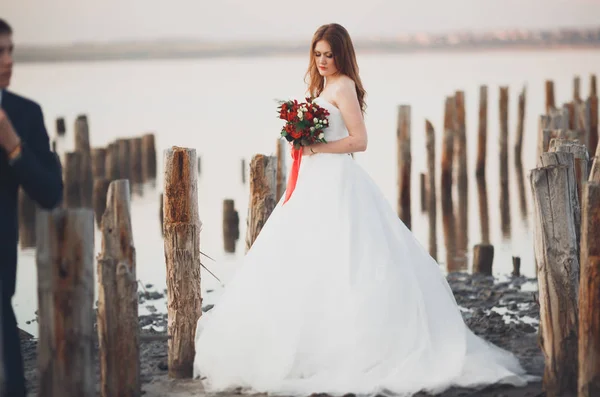 Hermosa joven pareja de boda, novia y novio posando cerca de postes de madera en el mar de fondo — Foto de Stock