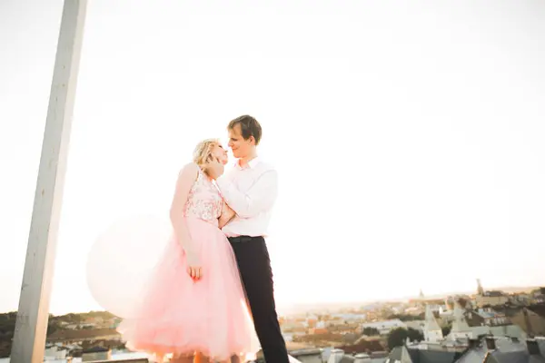 Pareja joven enamorada posando en el techo con una vista perfecta de la ciudad cogida de la mano y abrazándose. Hermoso atardecer — Foto de Stock