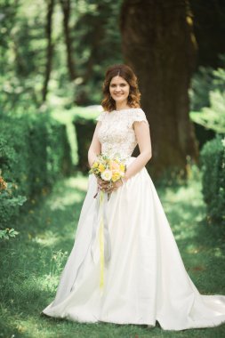 Zarif beyaz elbiseli güzel esmer gelin elinde buket ile güzel ağaçlar poz veriyor.