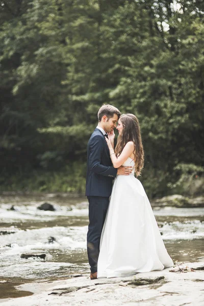 Elegante elegante novia morena feliz y hermoso novio en el fondo de un hermoso río en las montañas —  Fotos de Stock