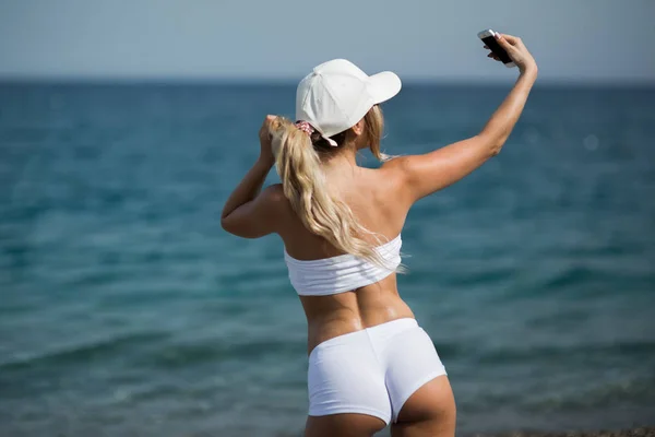 Beautiful blonde girl in sportwear on the beach makes selfie — Stock Photo, Image