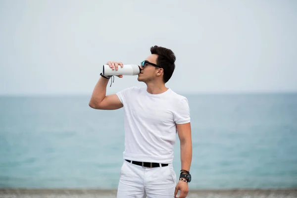 Joven en forma y deportivo está bebiendo de su botella de agua mientras disfruta de una carrera matutina en la playa — Foto de Stock