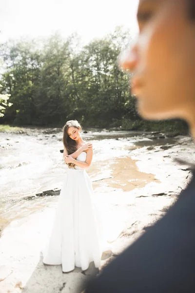 Pasangan pengantin yang cantik berciuman dan berpelukan di dekat tepi sungai pegunungan dengan batu — Stok Foto