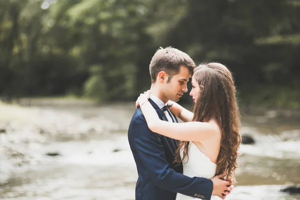 Elegan gaya pengantin berambut cokelat bahagia dan pengantin pria cantik di latar belakang sebuah sungai yang indah di pegunungan — Stok Foto