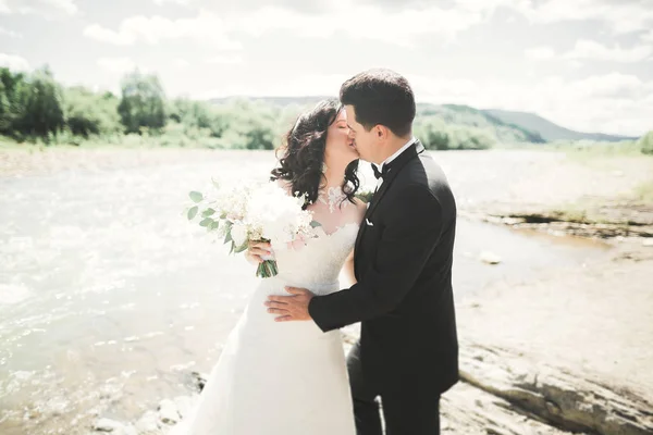 Hermosa pareja de boda besándose y abrazándose cerca de la orilla de un río de montaña con piedras —  Fotos de Stock