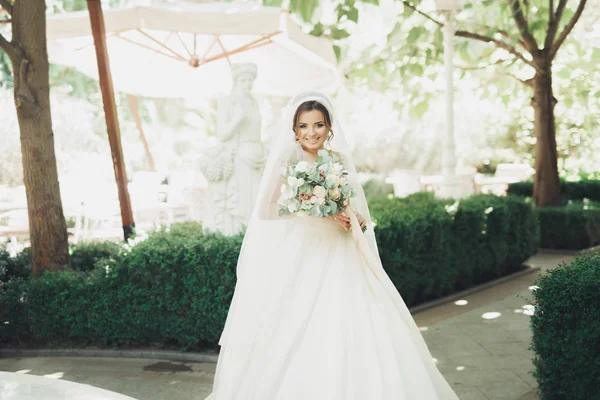 Hermosa novia morena en elegante vestido blanco sosteniendo ramo posando árboles limpios —  Fotos de Stock