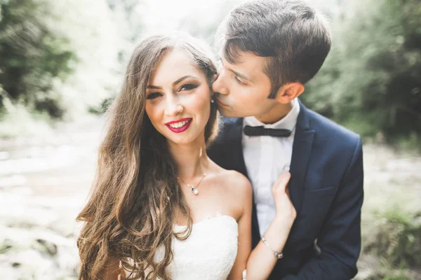 Hermosa pareja de boda besándose y abrazándose cerca de la orilla de un río de montaña con piedras — Foto de Stock