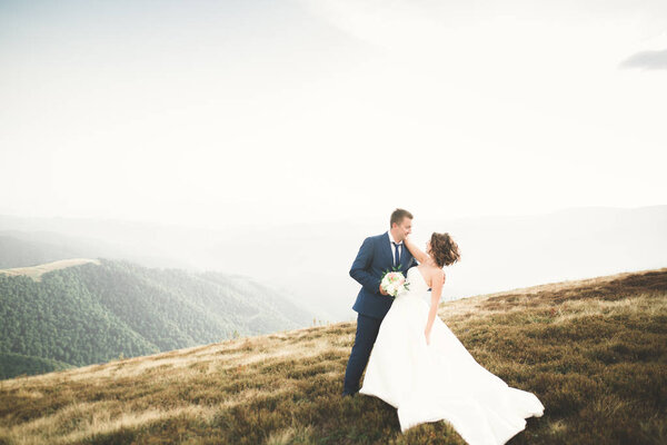 Young newly wed couple, bride and groom kissing, hugging on perfect view of mountains, blue sky.