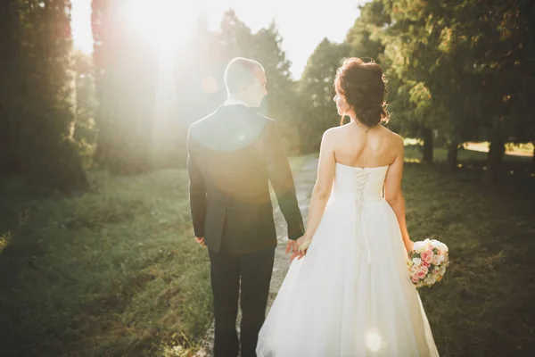 Elegante coppia di sposi felici che passeggiano nel parco il giorno del loro matrimonio con bouquet — Foto Stock