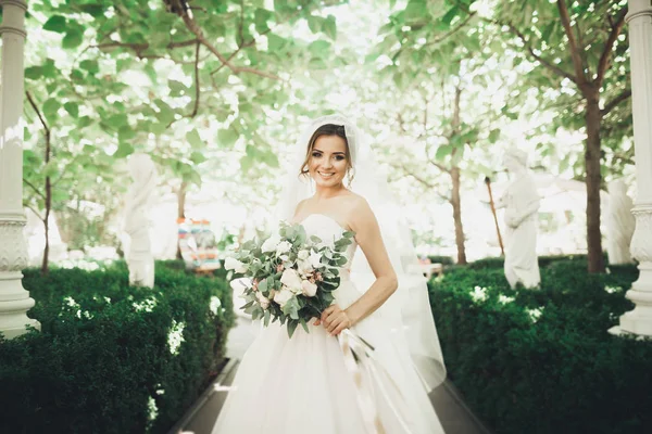 Mooie brunette bruid in elegante witte jurk met boeket poseren nette bomen — Stockfoto