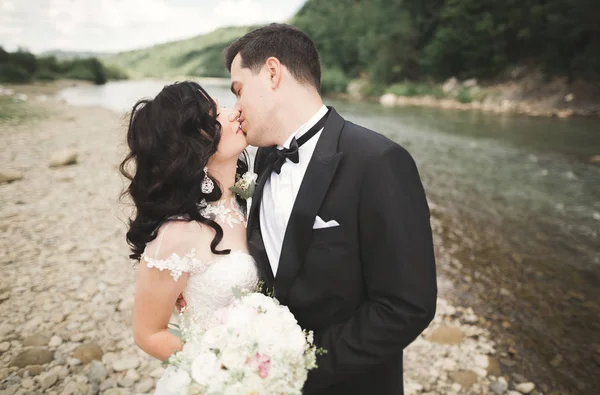 Schönes Hochzeitspaar küsst und umarmt sich am Ufer eines Gebirgsflusses mit Steinen — Stockfoto