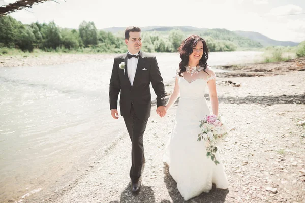 Hermosa pareja de boda besándose y abrazándose cerca de la orilla de un río de montaña con piedras —  Fotos de Stock