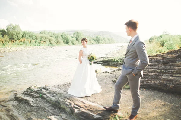 Guapo romántico novio y hermosa novia posando cerca del río en las montañas escénicas —  Fotos de Stock