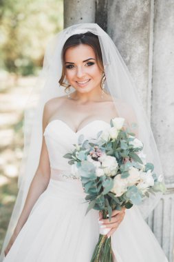 Zarif beyaz elbiseli güzel esmer gelin elinde buket ile güzel ağaçlar poz veriyor.