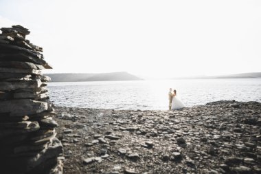 Düğün çifti mavi denizin yanındaki kayalarda öpüşüyor ve sarılıyor.