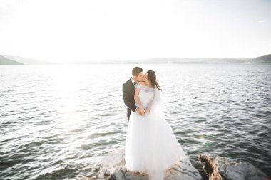 Düğün çifti mavi denizin yanındaki kayalarda öpüşüyor ve sarılıyor.