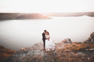 Düğün çifti mavi denizin yanındaki kayalarda öpüşüyor ve sarılıyor.