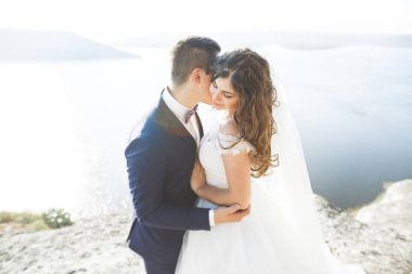 Düğün çifti mavi denizin yanındaki kayalarda öpüşüyor ve sarılıyor.