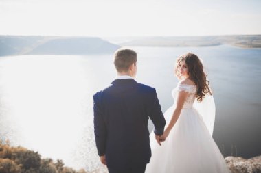 Düğün çifti mavi denizin yanındaki kayalarda öpüşüyor ve sarılıyor.