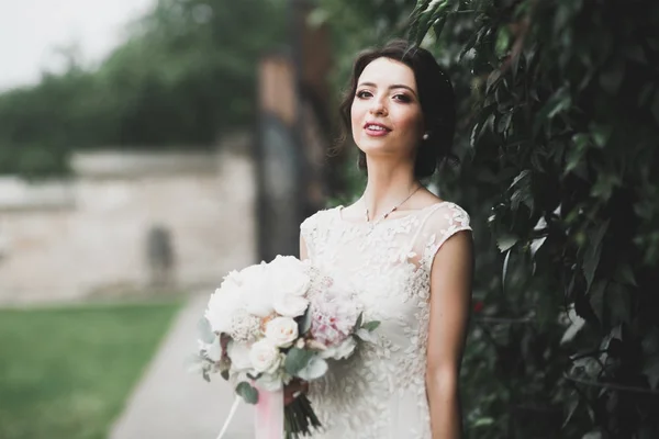 Bella sposa bruna in elegante vestito bianco che tiene bouquet in posa alberi ordinati — Foto Stock