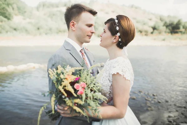 Hermosa pareja de boda besándose y abrazándose cerca de la orilla de un río de montaña con piedras —  Fotos de Stock