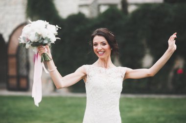 Zarif beyaz elbiseli güzel esmer gelin elinde buket ile güzel ağaçlar poz veriyor.