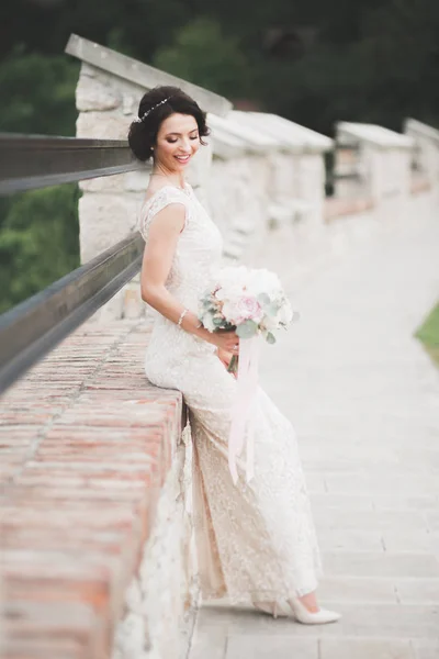 Bela noiva morena em vestido branco elegante segurando buquê posando árvores limpas — Fotografia de Stock