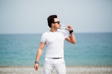 Zinde ve sportif görünen genç adam sahilde koşmak bir sabah tadını çıkarırken onun su şişesinden içiyor