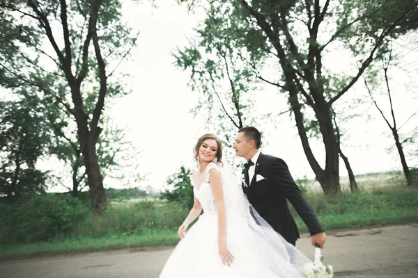 Стильная пара счастливых молодоженов, прогуливающихся в парке в день своей свадьбы с букетом — стоковое фото