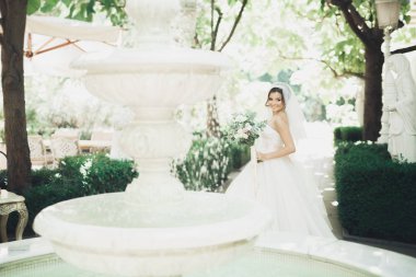 Zarif beyaz elbiseli güzel esmer gelin elinde buket ile güzel ağaçlar poz veriyor.