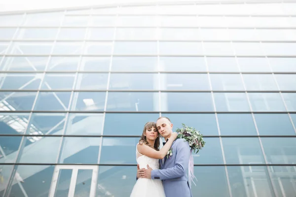 Casal feliz encantador do casamento, noiva com vestido branco longo posando perto do edifício moderno — Fotografia de Stock