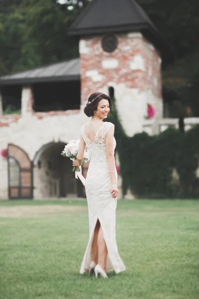 Hermosa novia morena en elegante vestido blanco sosteniendo ramo posando árboles limpios — Foto de Stock