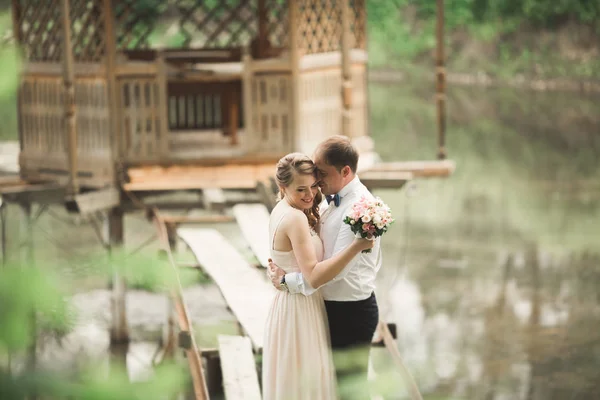 Прекрасная влюбленная пара целует друг друга в день свадьбы, стоя в парке на открытом воздухе возле озера — стоковое фото
