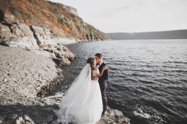 Düğün çifti mavi denizin yanındaki kayalarda öpüşüyor ve sarılıyor.