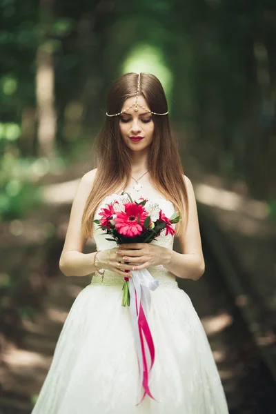 Bella sposa bruna in elegante vestito bianco che tiene bouquet in posa alberi ordinati — Foto Stock