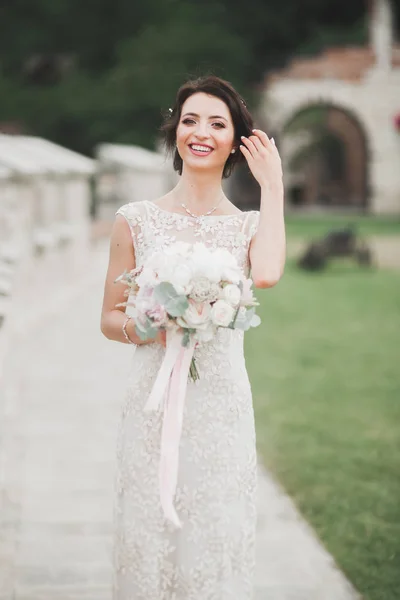 Bella sposa bruna in elegante vestito bianco che tiene bouquet in posa alberi ordinati — Foto Stock