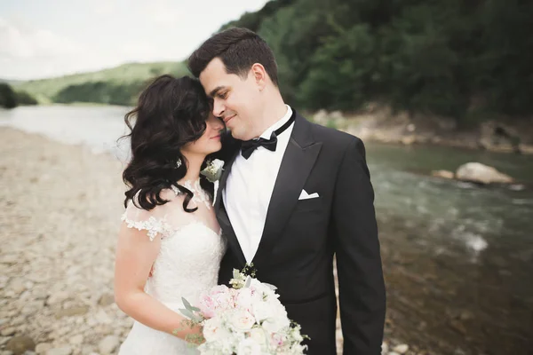 Elegante stilvolle glückliche brünette Braut und wunderschöner Bräutigam vor dem Hintergrund eines wunderschönen Wasserfalls in den Bergen — Stockfoto