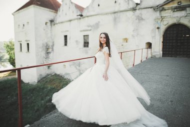 Zarif beyaz elbiseli güzel esmer gelin elinde buket ile güzel ağaçlar poz veriyor.